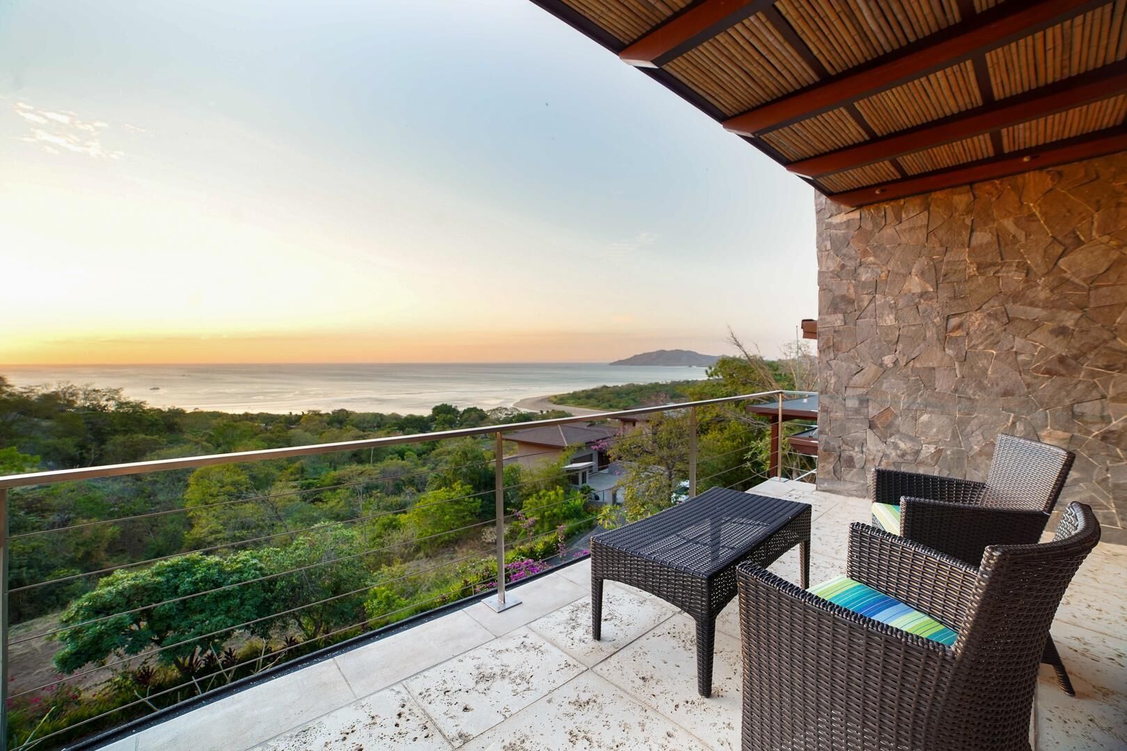A balcony with wicker furniture, a stone wall, and a view of the ocean and hills.