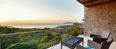 A balcony with wicker furniture, a stone wall, and a view of the ocean and hills.