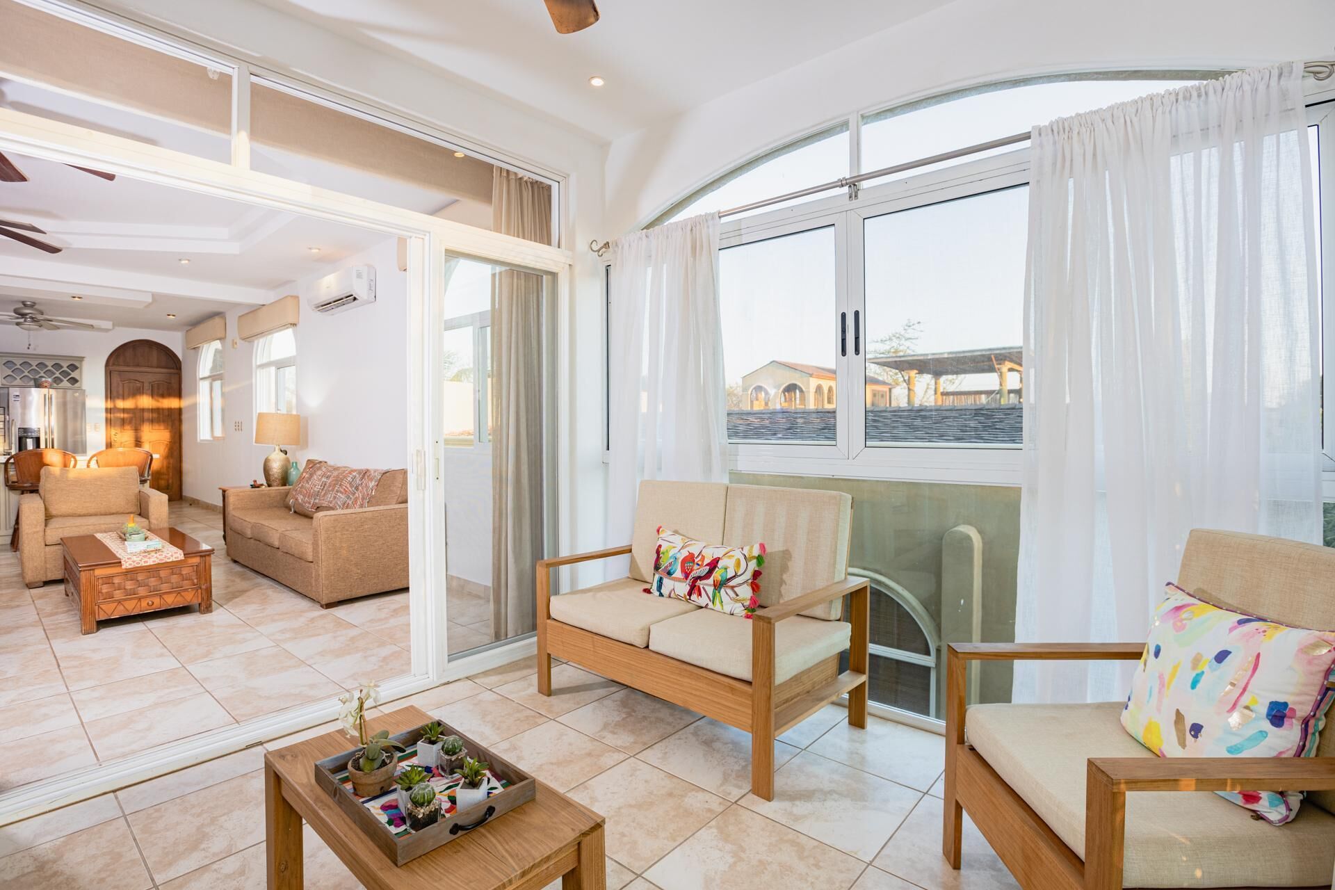 A spacious living area with a sofa, armchairs, and a coffee table. There is a sliding glass door leading to a balcony with a view of buildings.