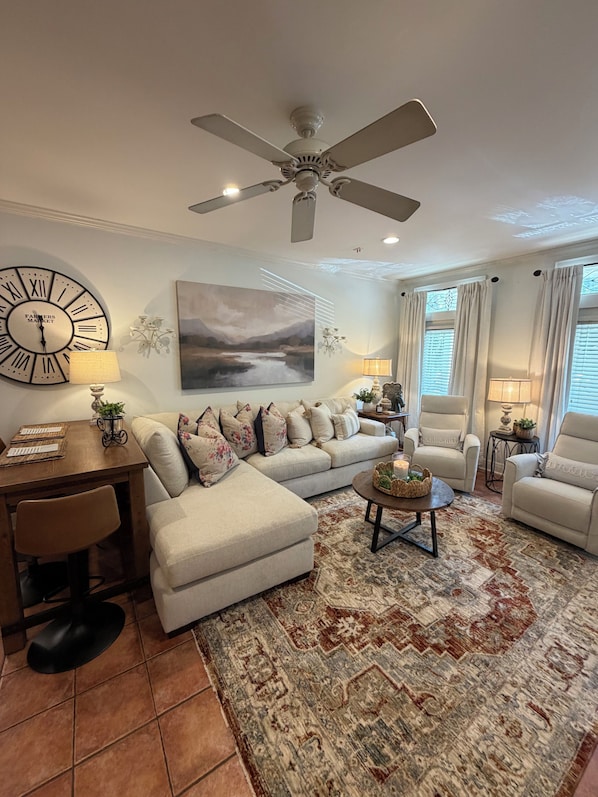 Un salon avec un ventilateur de plafond, une grande horloge, un canapé, une table basse et un tapis.