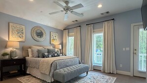 Une chambre à coucher avec un lit, un ventilateur de plafond, une grande fenêtre avec des rideaux, un tapis et des œuvres d’art murales.