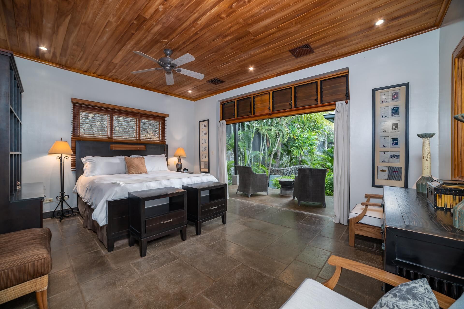 A bedroom with a bed, bedside tables, a desk, and a chair. There is a window with blinds and a view of greenery outside.
