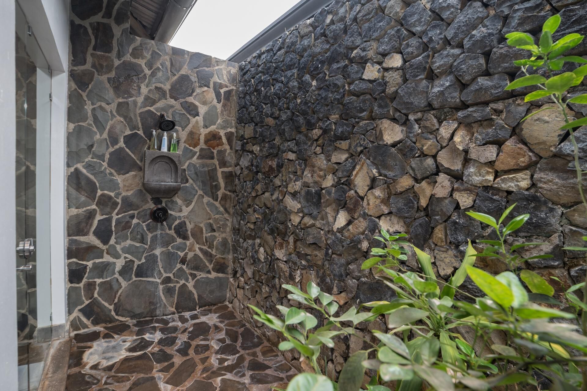 A stone-walled outdoor area with a built-in shower and a small shelf holding toiletries.