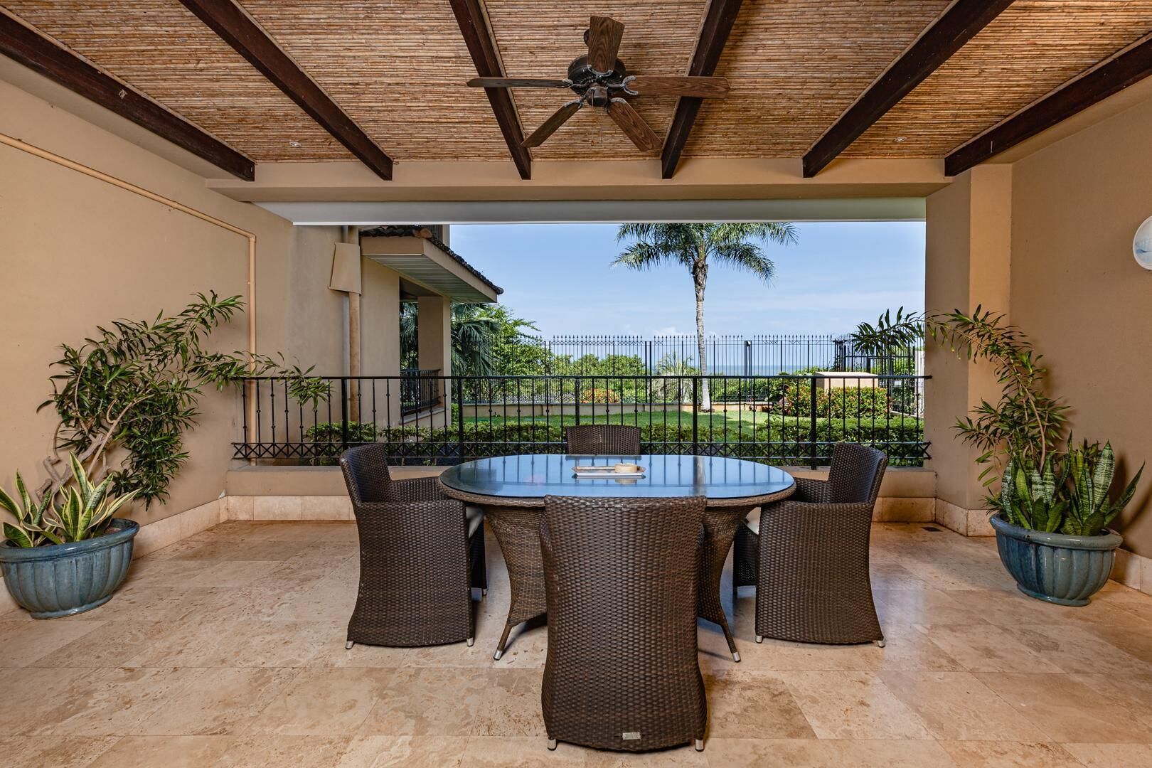 A covered patio with a round table and wicker chairs, surrounded by potted plants, and overlooking a landscaped garden.