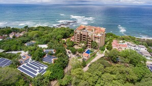 Et luftbillede af et kysthotel med flere bygninger, herunder en markant bygning med flere etager og rødt tegltag. Solpaneler er synlige på taget, og hotellet er omgivet af frodig vegetation og havet.