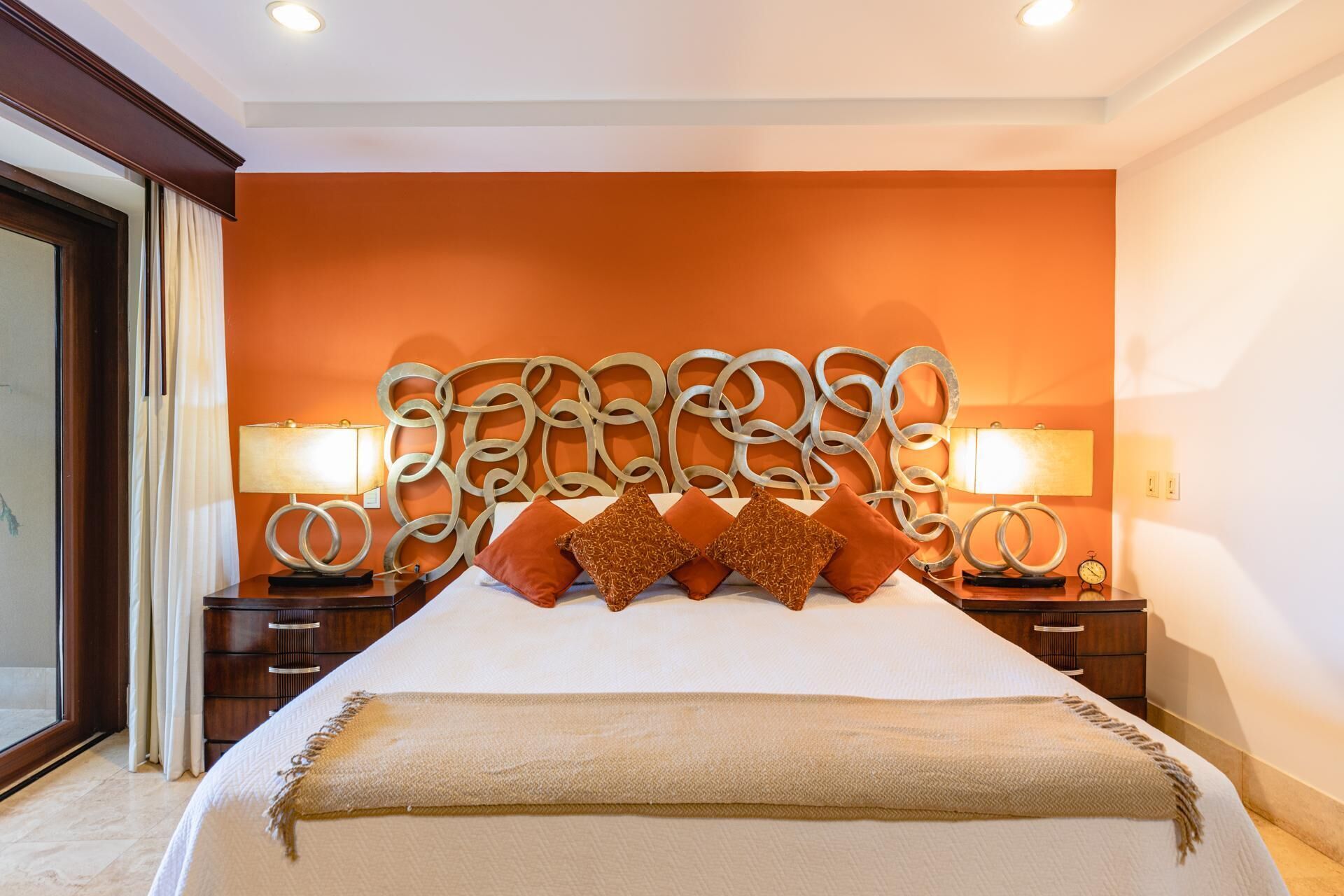 A hotel room with a large bed, two bedside tables with lamps, an orange accent wall, and a view through a large window.