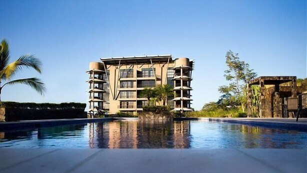 A modern hotel with multiple floors and balconies, a swimming pool in the foreground, and tropical landscaping.