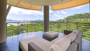 A balcony with a view of a beach and hills, furnished with wicker furniture and a cushioned bench.