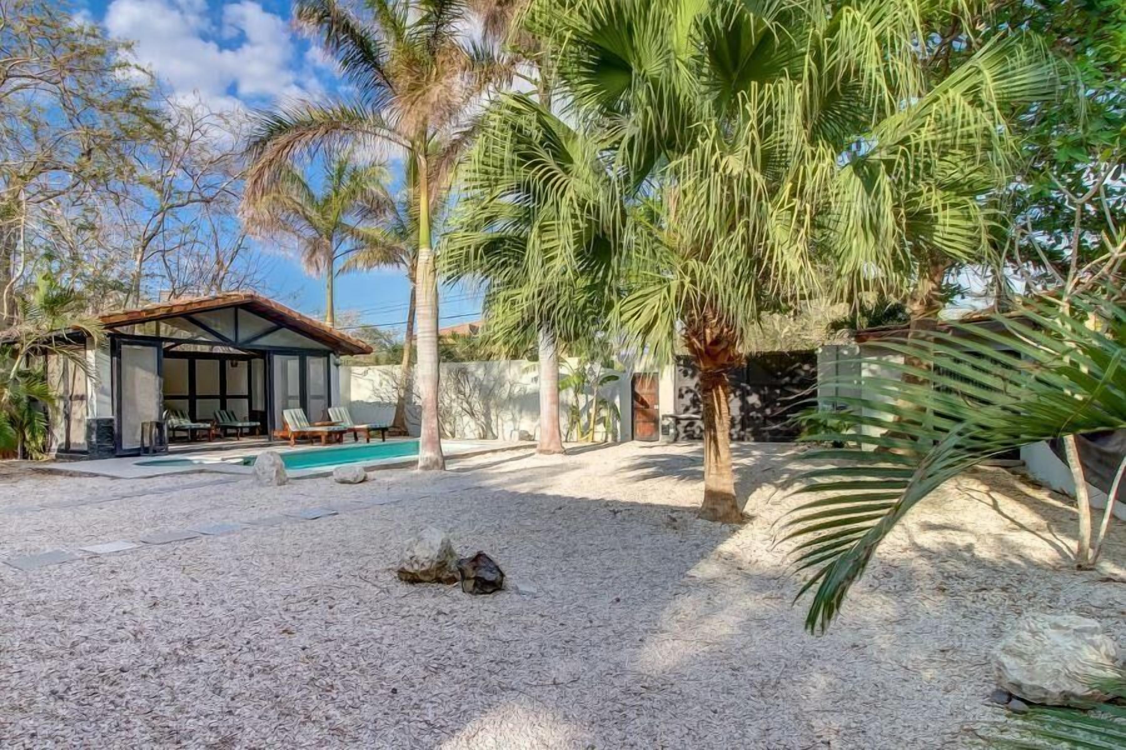 A covered outdoor area with a pool, palm trees, and a building with a tiled roof.