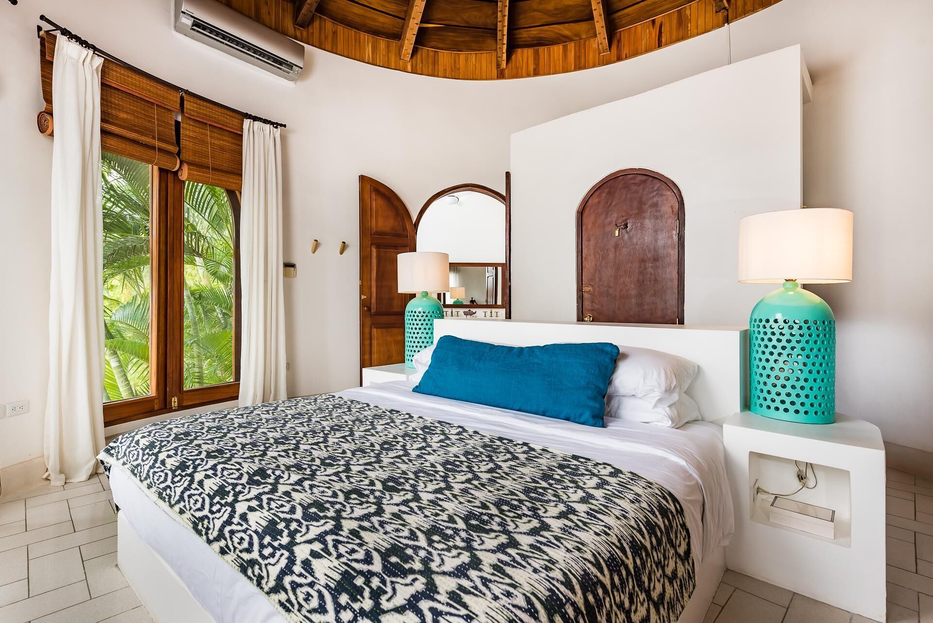 A bedroom with a patterned bedspread, two bedside lamps, a wooden door, and a large window with curtains.