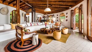 A spacious living area with a high wooden ceiling, a white sofa with colorful cushions, a wooden coffee table, and a large window offering a view of greenery. - Flamingo Blu (Cabo Velas)