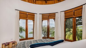 A bedroom with a large bed, a wooden bench, and three windows with wooden frames and white curtains. - Flamingo Blu (Cabo Velas)