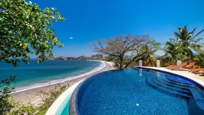 A clear blue pool with a curved edge, surrounded by lounge chairs and palm trees, overlooking a beach and ocean. - Flamingo Blu (Cabo Velas)