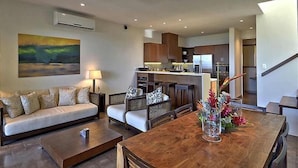 Una sala moderna con sofá, una mesita de centro y una mesa de comedor con un jarrón de flores. Se observa una zona de cocina con armarios de madera y un refrigerador.