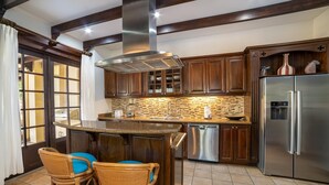 Eine moderne Küche mit dunklen Holzschränken, einer Edelstahl-Dunstabzugshaube, einem eingebauten Geschirrspüler und einem Kühlschrank mit Wasserspender. Zwei Korbstühle mit blauen Kissen stehen an einer kleinen Kochinsel.