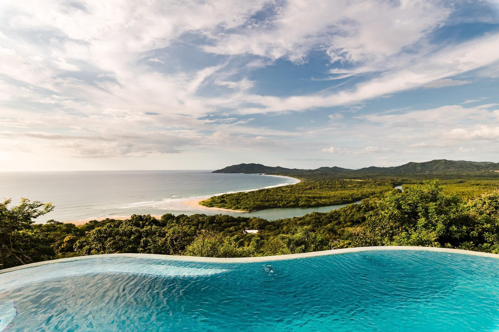 A clear view of a swimming pool with a curved edge, surrounded by lush greenery and overlooking a beach with a curved shoreline.