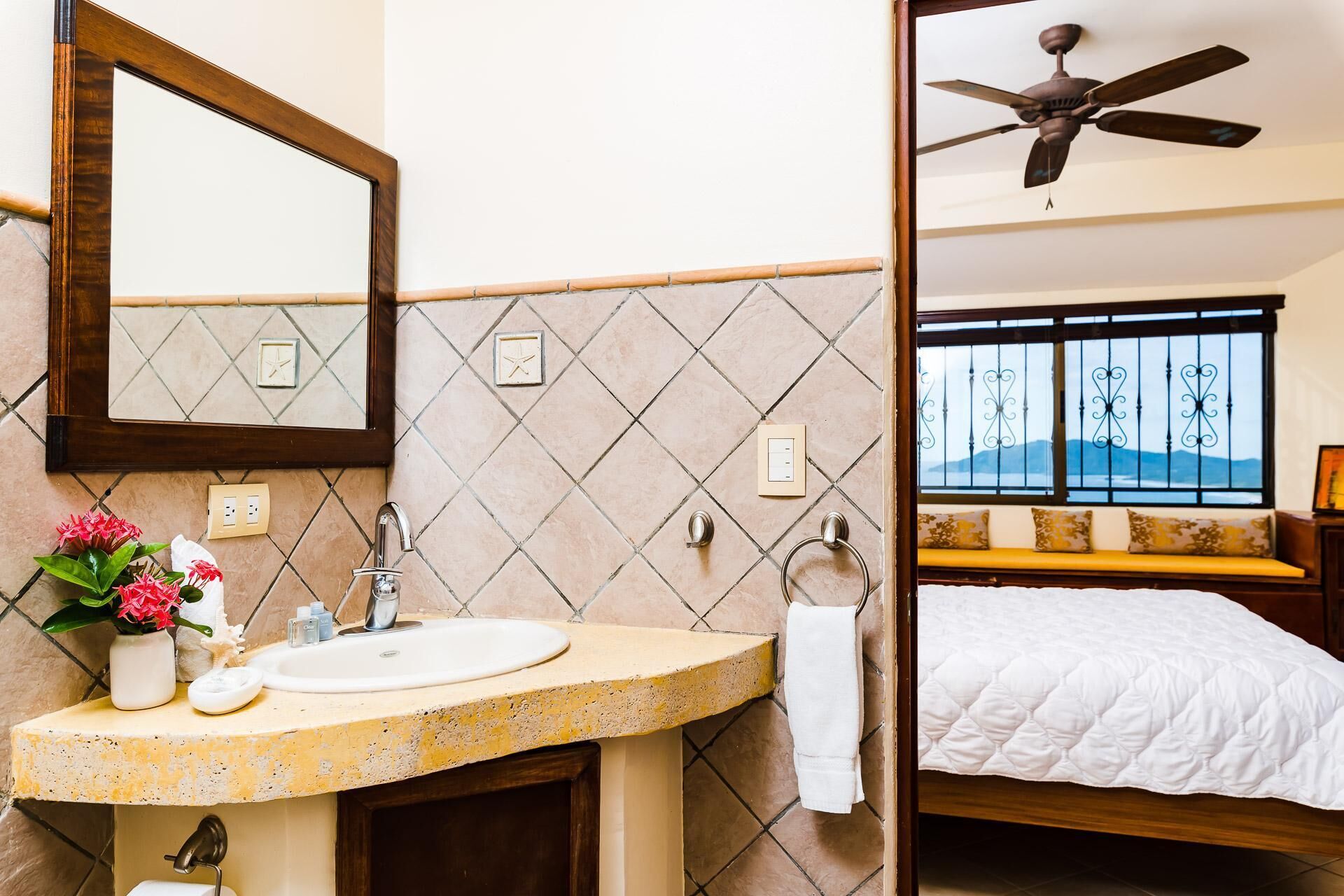 Baño con lavamanos, espejo y flores decorativas. Incluye también una cama con vistas a las montañas a través de la ventana.