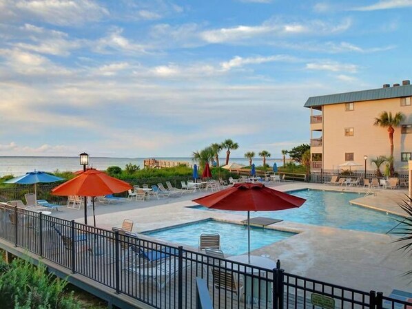A pool area with lounge chairs and umbrellas, a view of the ocean, and a building with balconies.