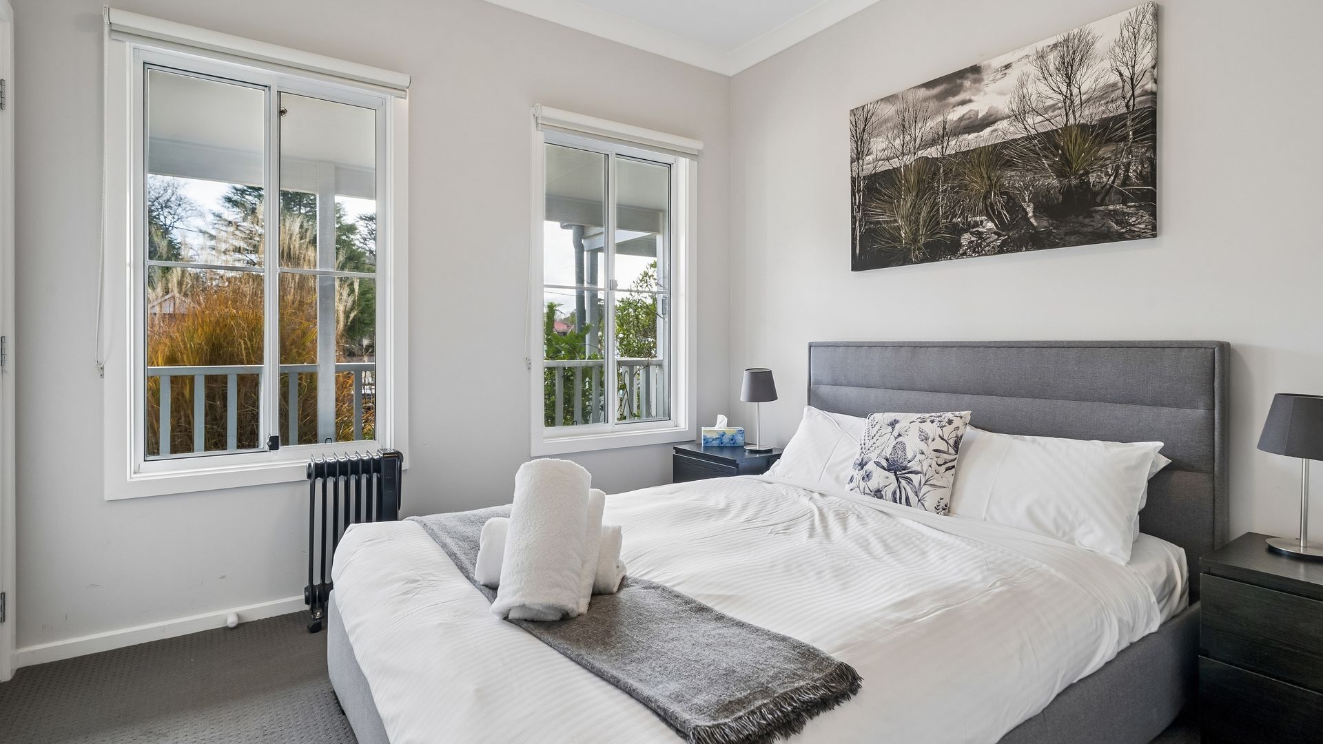 A bedroom with a large bed, two windows, a radiator, a bedside table with a lamp, and a wall-mounted picture.