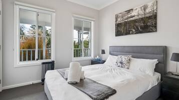A bedroom with a large bed, two windows, a radiator, a bedside table with a lamp, and a wall-mounted picture.