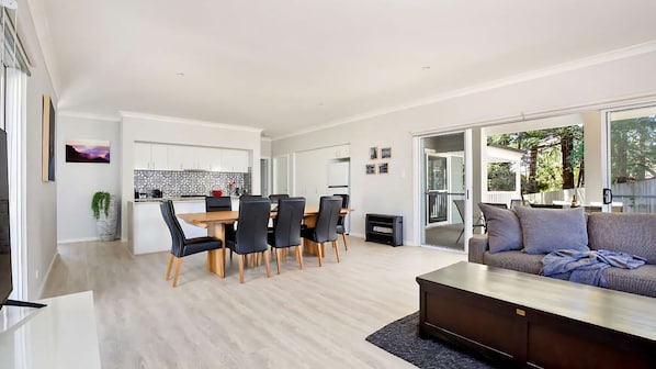 Un salon moderne avec un canapé et une table basse, un coin repas avec une table et des chaises, et une cuisine avec des meubles blancs et un crédence.