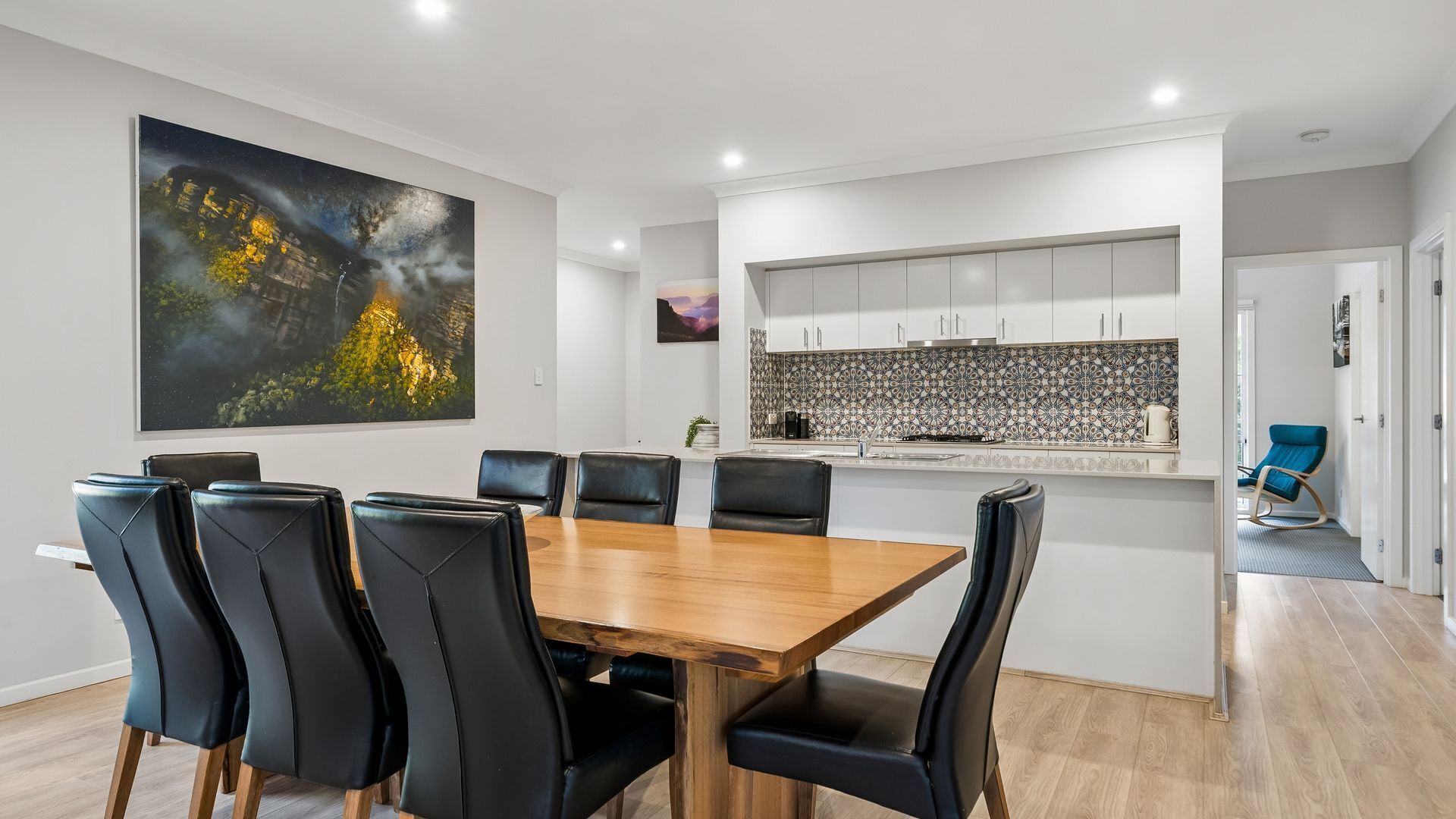 A modern dining area with a wooden table and black chairs, a kitchen with patterned backsplash, and a large abstract painting on the wall.