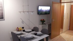 Un área de comedor con una mesa preparada para cuatro personas, un televisor montado en la pared y una puerta que da a otra habitación.
