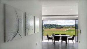 Ein moderner Essbereich mit großem Tisch und Stühlen, Blick auf eine ländliche Landschaft durch eine Glastür und abstrakte Wandkunst.