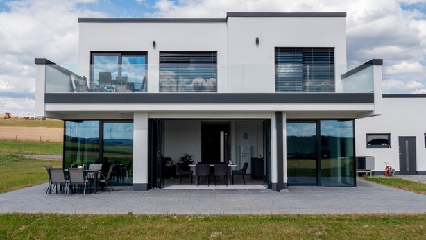 Ein modernes, zweistöckiges Haus mit Balkon, großen Fenstern und einer gepflasterten Terrasse.