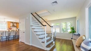 Amplia sala de estar con pisos de madera, una escalera y un área de cocina con gabinetes de madera y una barra con asientos.