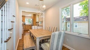 Un comedor con una mesa de madera y sillas a rayas, una cocina al fondo y una ventana con vista a una casa.