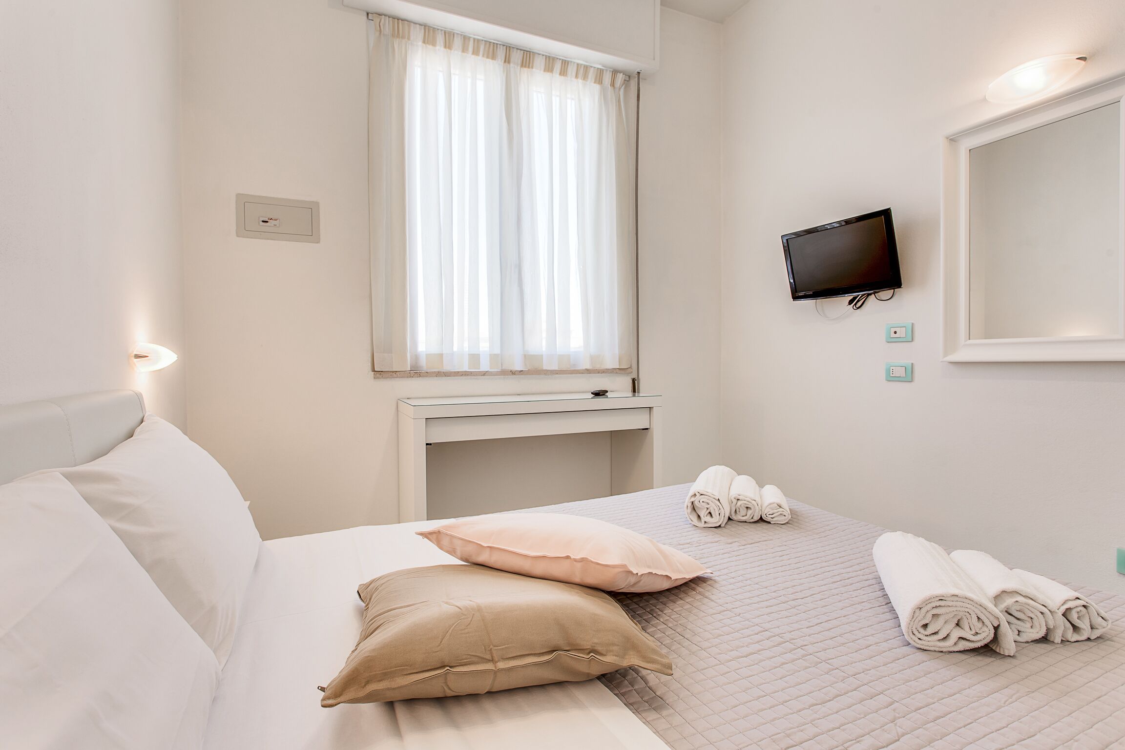 A hotel room with a bed, pillows, a TV, a desk, a window with curtains, and rolled towels on the floor.