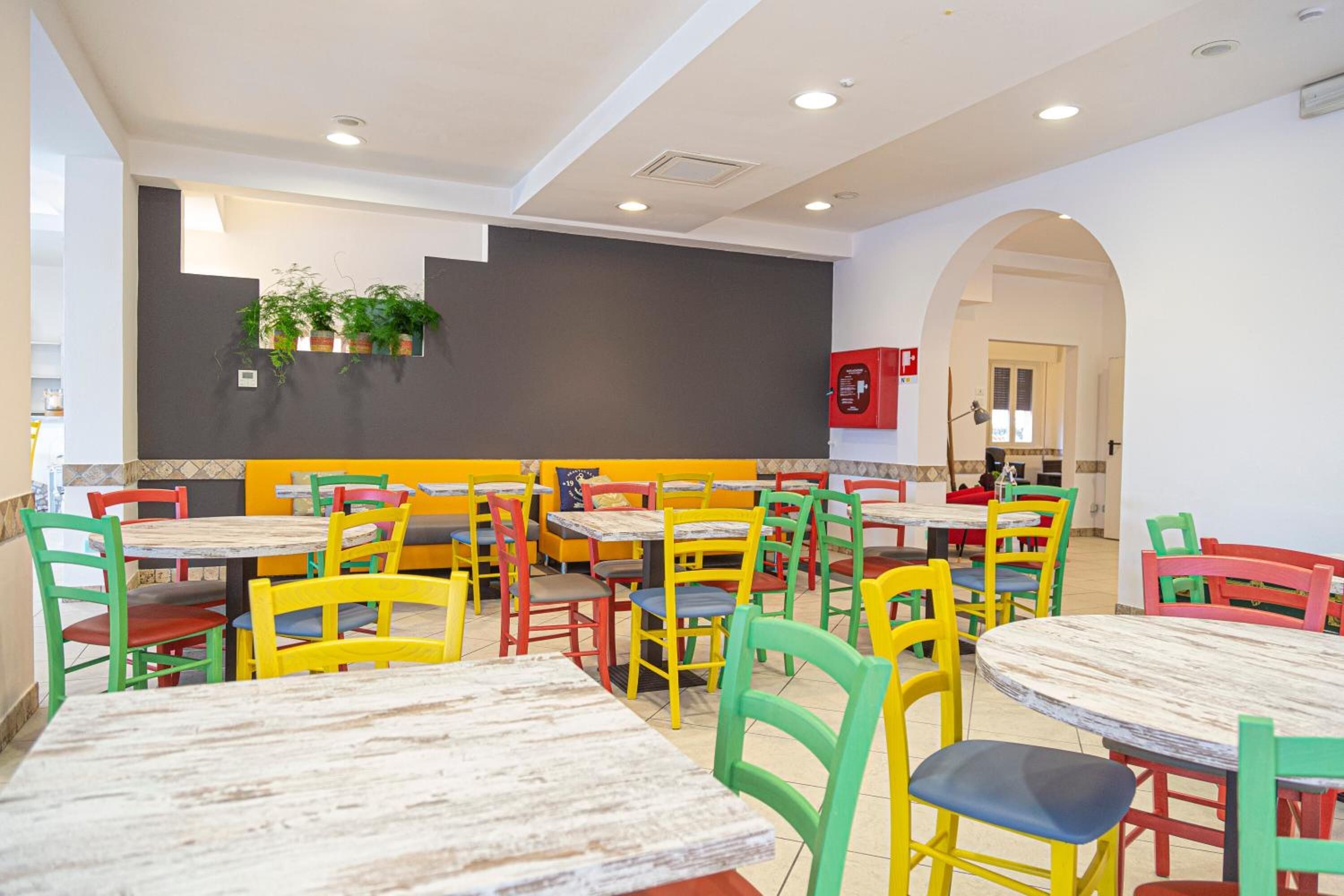 A restaurant with multiple tables and colorful chairs, a grey wall with plants, and a red fire extinguisher.