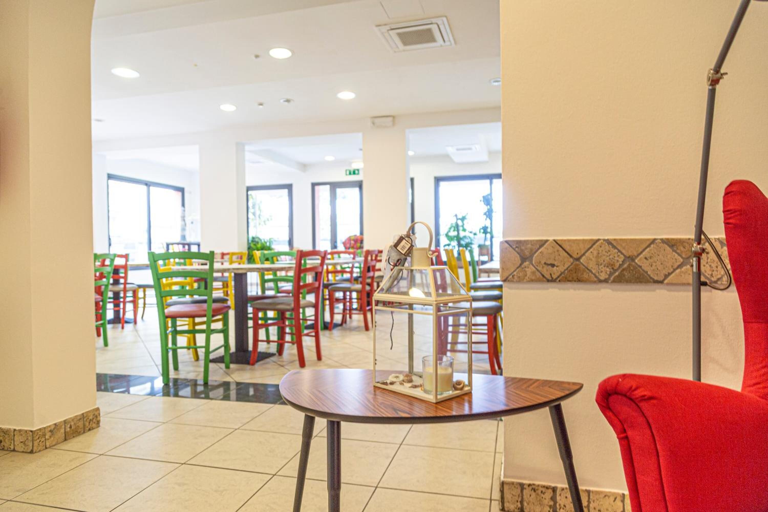 Un vestíbulo de hotel con sillas coloridas, una mesa con un farol y ventanas amplias.