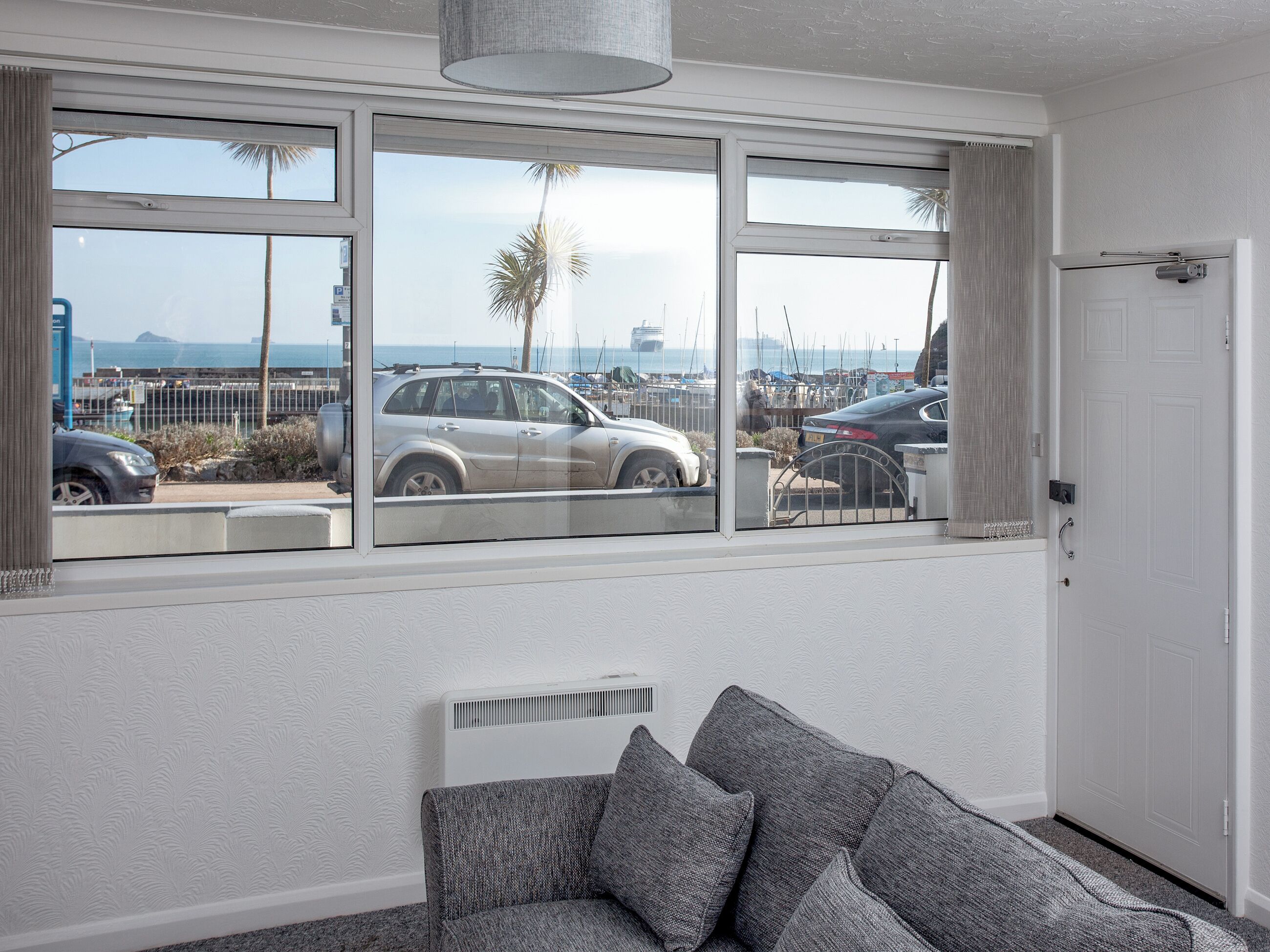 Una habitación con un gran ventanal con vista a un puerto deportivo, una puerta blanca y un sofá gris.