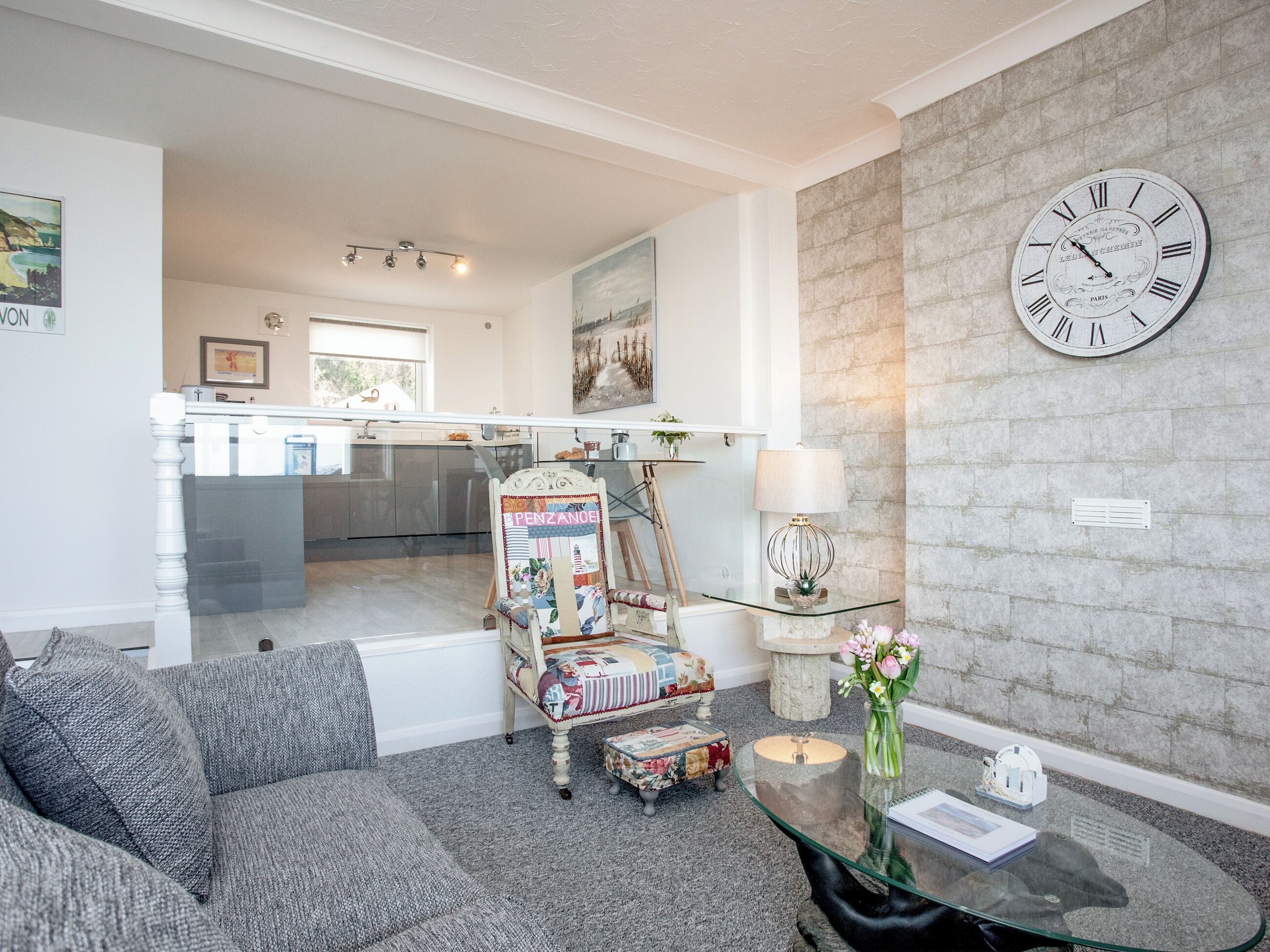 Een woonkamer met een glazen salontafel, een bank, een klok aan de muur en een keuken op de achtergrond.
