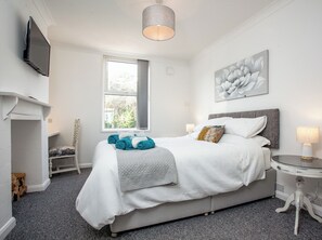 Un dormitorio ordenado con cama, mesita de noche, sillón, chimenea, ventana y un cuadro en la pared.