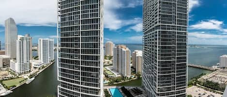 A high-rise building with a glass facade, surrounded by other high-rise buildings, a body of water, and a clear sky.