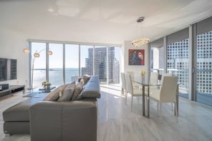 A modern living room with a large sofa, dining table, and chairs, large windows with a city view, and a mounted TV.