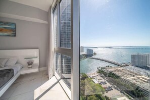 A modern bedroom with a large window, a bed, a bedside table, and a view of a coastal cityscape.