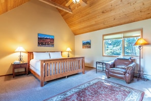Quarto com cama de madeira, duas luminárias de cabeceira, poltrona de couro e uma janela grande com vista para árvores.