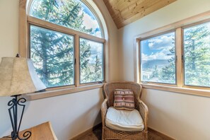 A corner room with two large windows offering a view of trees and mountains. A wicker chair with a cushion is placed against the wall.