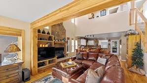 A spacious living area with a large brown leather sofa, wooden beams, a stone fireplace, and a kitchen in the background.