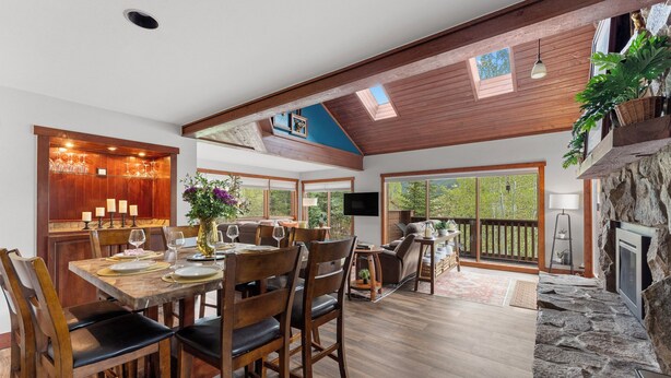 A spacious dining area with wooden tables and chairs, a stone fireplace, and large windows offering views of the outdoors. - 67 W Hanson Road by Summit County Mountain Retreats (Keystone)