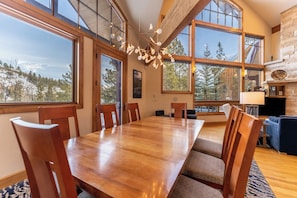 A dining area with a wooden table and chairs, large windows, and a stone fireplace. - 580 Elk Circle by Summit County Mountain Retreats (Keystone)