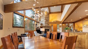 A spacious dining area with a long wooden table and chairs, a stone fireplace, and large windows offering a view of trees.