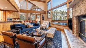 A spacious living area with a fireplace, large windows, and a kitchen in the background.