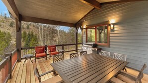 A covered deck with a wooden table and chairs, a grill, and a view of the surrounding landscape.