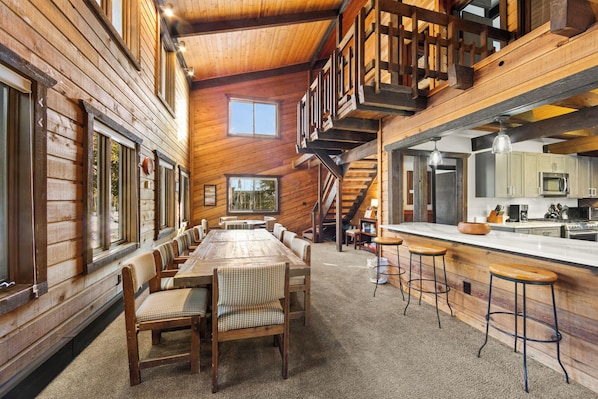 A spacious wooden cabin with a long dining table, multiple bar stools, a kitchen area, and a staircase leading to an upper level.