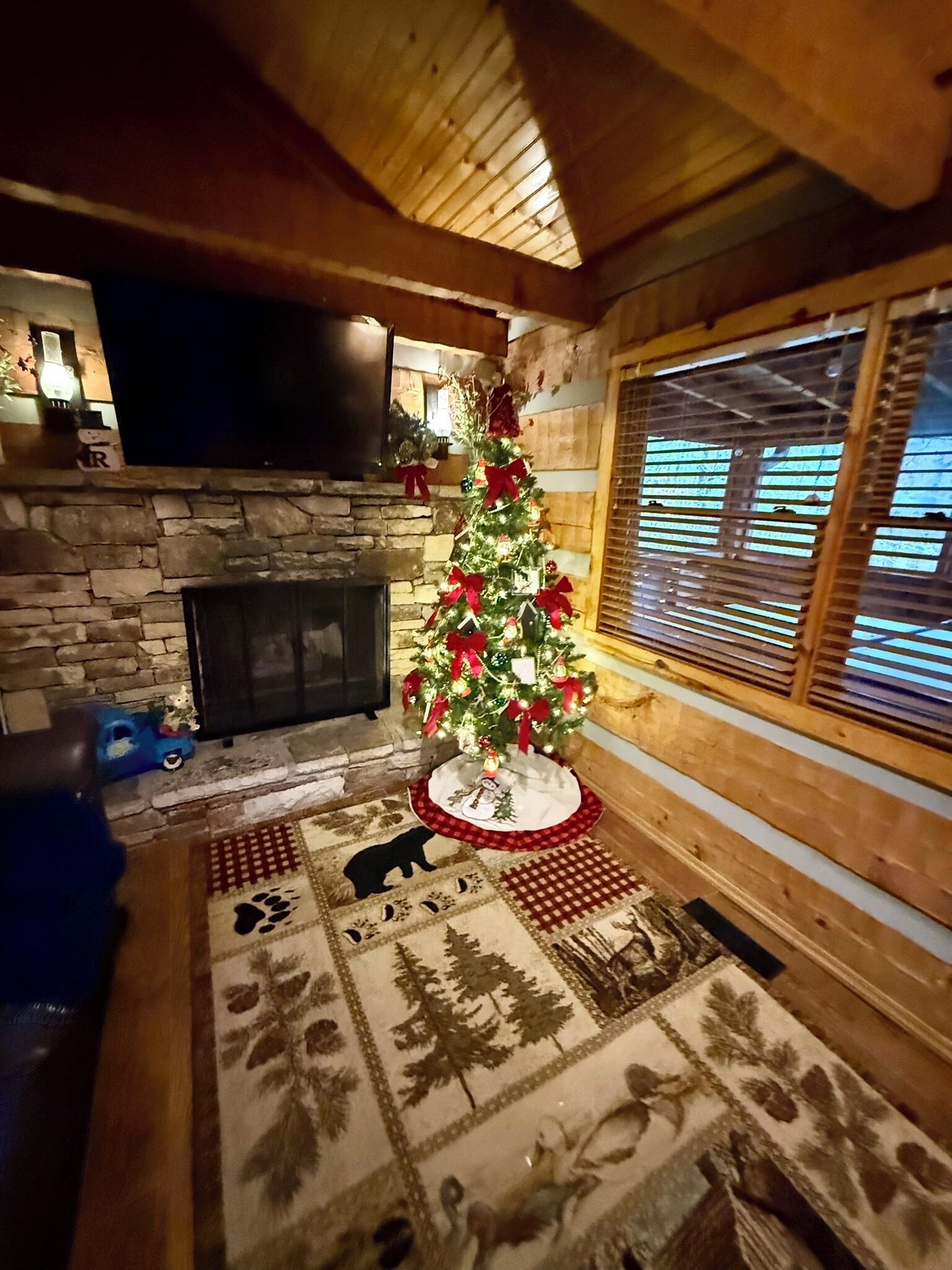 A cozy room with a stone fireplace, a Christmas tree, and a large window with blinds.
